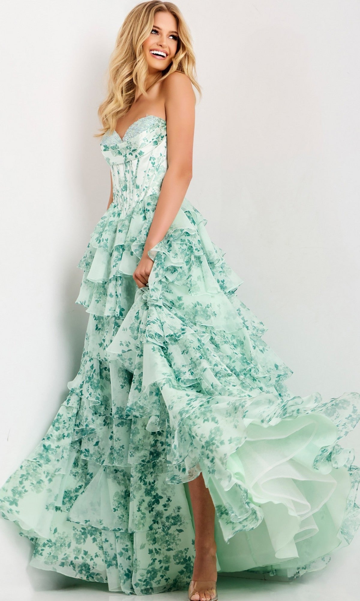 Woman models the Jovani 48772 long prom dress, a strapless ball gown with a mint green floral print and ruffled skirt, posing against a white background while lifting part of the skirt and smiling.